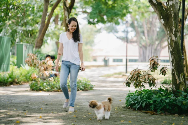 Portrait of a young Pan Asian woman sitting with her young shih tzuh puppy dog in the park in the day. She is smiling and they are both enjoying the warm day.