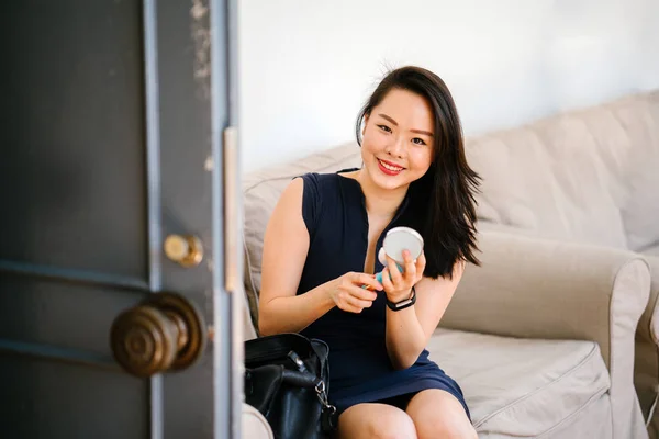 young Chinese Asian woman is applying makeup on a couch in the day next to a door. She\'s smiling and is looking cheerful, sunny and is professionally dressed in a dark, navy blue dress with bag