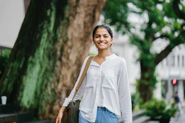 魅力的で若いインドの女性は日に都市の木の隣に立っています 彼女はカジュアルな白いブラウスとジーンズです 植民地時代の古い建物とバック グラウンドでのトラフィックがあります — ストック写真