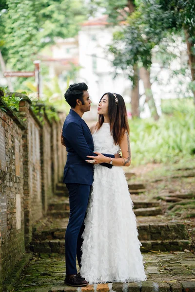 interracial couple (Indian man, Chinese woman) pose for wedding photo in a park in the day. They are on old stone steps and and smiling and laughing as they take photos.