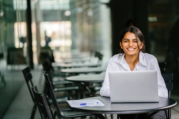 young Indian Asian woman is typing and working in a laptop in the day (applying for a job, etc). She is wearing a professional, crisp white shirt and she is smiling as she types on her laptop.