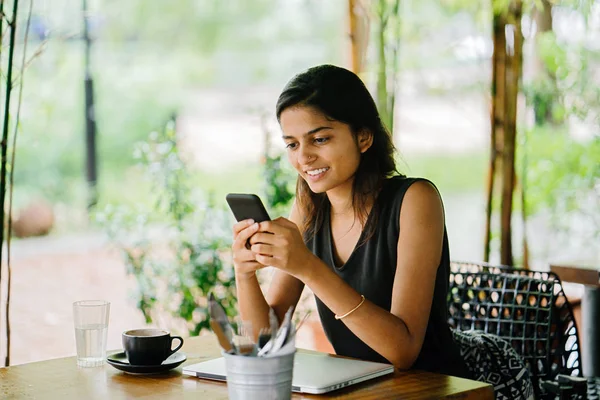 一个迷人的 年轻的印度妇女坐在咖啡馆或 Coworking 空间与智能手机的坦率肖像 在一个晴朗的日子 有茂盛的 绿色的植物在背景 — 图库照片