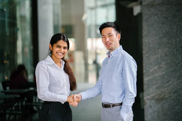 Two professional businesspeople shake hands in an office in the city. An Indian woman shakes hands with a Chinese man in agreement and they are both smiling and professionally dressed.