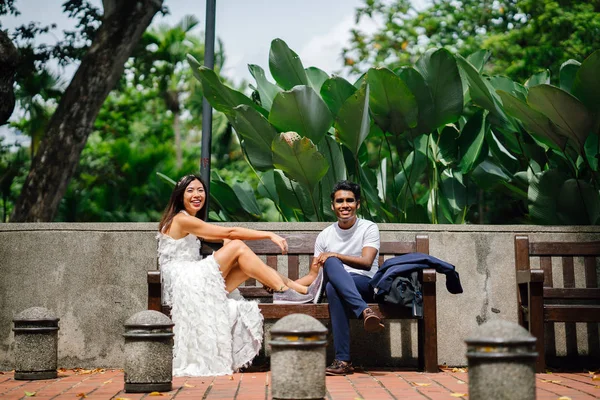 bride and groom share a relaxed moment on a bench in the park. They are an interracial couple from different cultures, one is an Indian man the other a Chinese woman.