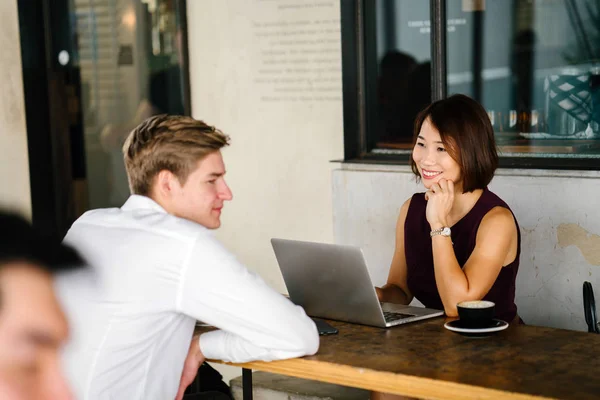 An Asian Chinese woman interviews a young, Caucasian man. She is looking a her laptop computer as she talks with him and they are both smiling. They are sitting at a table in the day in an office.