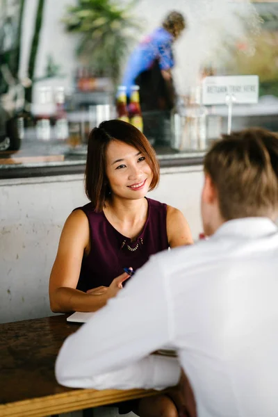 An Asian woman is interviewing a young Caucasian white man for an internship job in Asia. They are sitting at a desk in the shade outside in the day and talking over coffee.