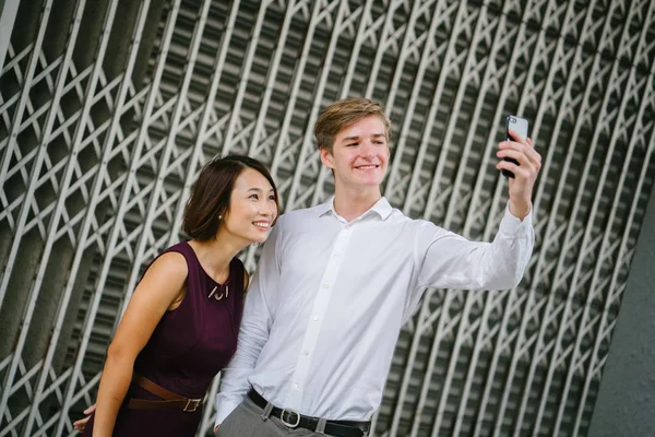 mixed-culture couple take a selfie in the day against the metal grilles of a closed Chinese shop in Asia. A young and attractive white man smiles next to a Chinese woman as they take their photo.