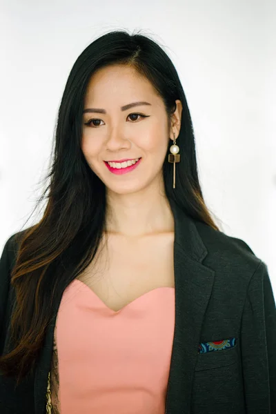 Portrait of a young Asian Chinese woman (blogger, influencer, fashionista) standing against a white plain background. She is very fashionably dressed and has a man's jacket draped on her shoulders.
