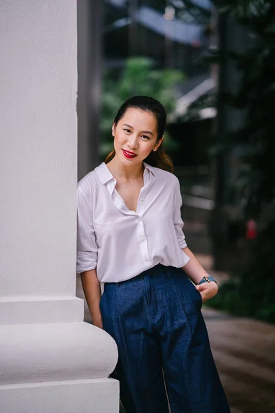 A young Singaporean Chinese woman lounges and relaxes against the wall in the city in Singapore during the day. She is professionally and elegantly dressed in a white shirt and navy pants.