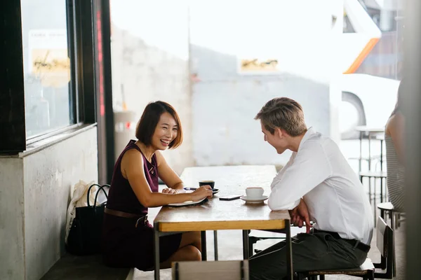 An Asian woman is interviewing a young Caucasian white man for an internship job in Asia. They are sitting at a desk in the shade outside in the day and talking over coffee.