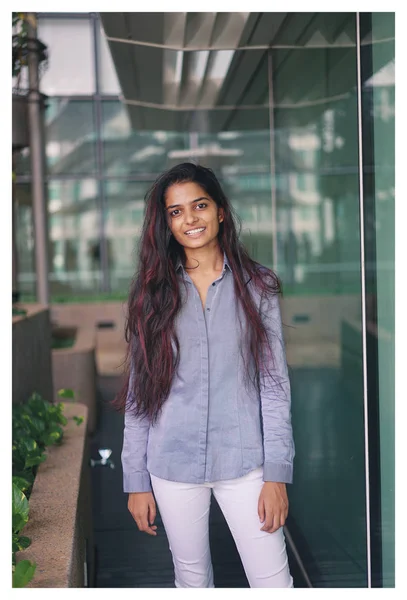 Portrait of a young Indian woman standing near the glass wall wearing formal clothing. She is casually smiling back towards the camera in a natural way.