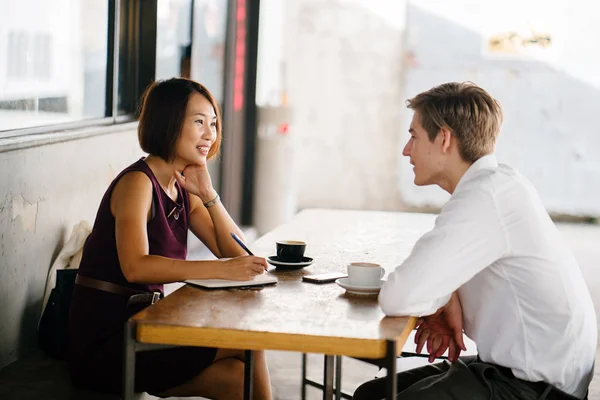 An Asian woman is interviewing a young Caucasian white man for an internship job in Asia. They are sitting at a desk in the shade outside in the day and talking over coffee.