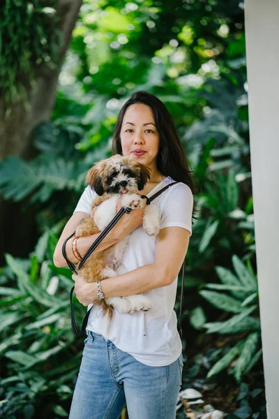 Portrait of a young Pan Asian woman carrying her dog and smiling for the camera in a green park on a warm day in the park. The dog is a toy breed shih tzuh.
