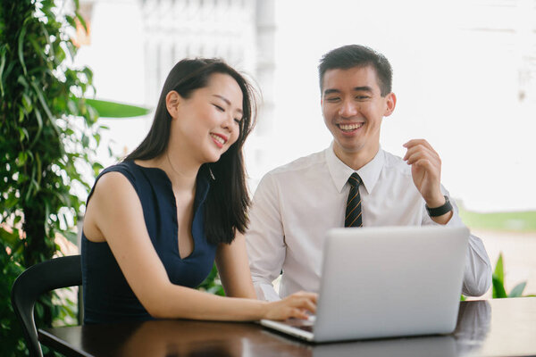 Portrait of two smiling business people (lawyers, consultants, etc) or couple smiling mid discussion over a laptop in an office with greenery in the background. They are focused on the laptop.