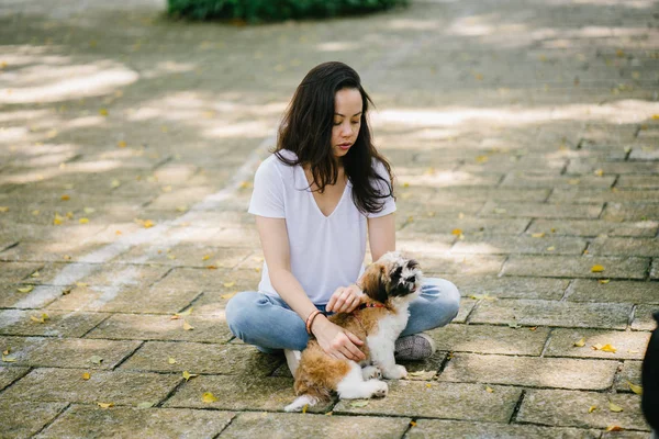 Portrait of a young Pan Asian woman sitting with her young shih tzuh puppy dog in the park in the day. She is smiling and they are both enjoying the warm day.
