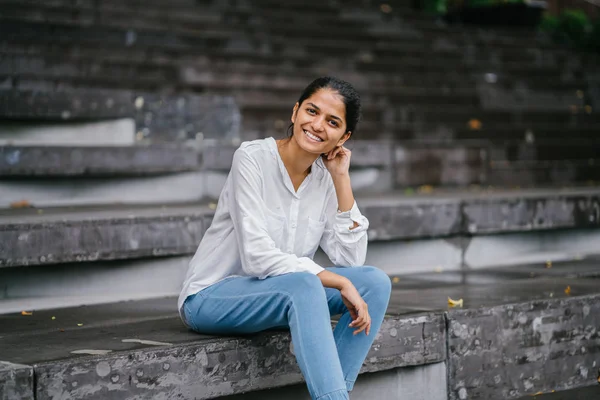 Candid portrait of a smiling attractive, young woman on grey steps . The Indian Asian lady is professional, relaxed and sitting in the bright sun.