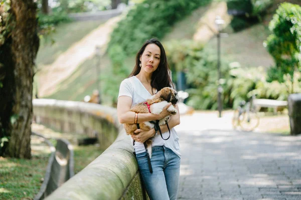 Portrait of a young, attractive pan Asian woman with her shih tzuh dog in the park. They are in the middle of a walk and they look cheerful and relaxed.