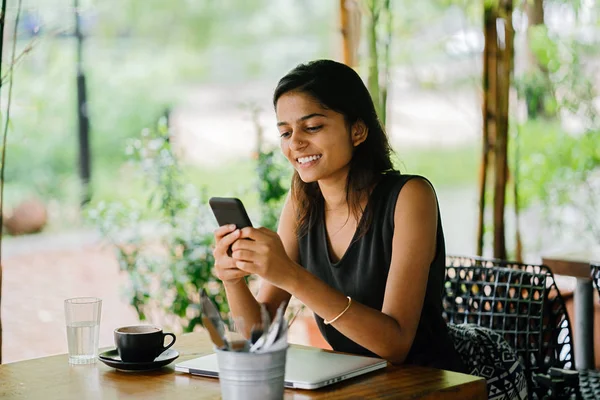 一个迷人的 年轻的印度妇女坐在咖啡馆或 Coworking 空间与智能手机的坦率肖像 在一个晴朗的日子 有茂盛的 绿色的植物在背景 — 图库照片