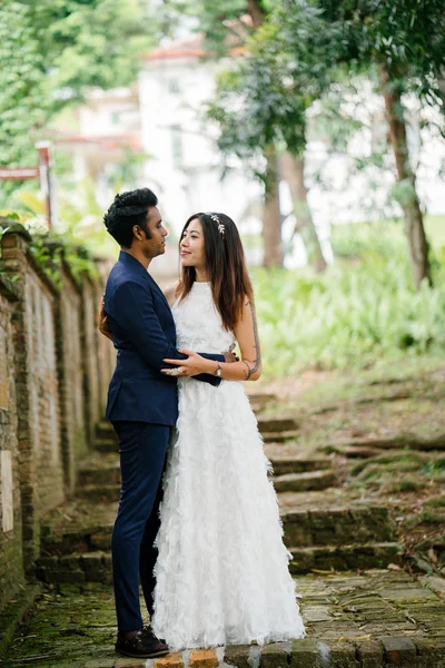 interracial couple (Indian man, Chinese woman) pose for wedding photo in a park in the day. They are on old stone steps and and smiling and laughing as they take photos.