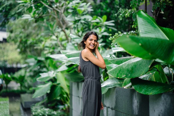 Candid portrait of a young Indian woman standing in a lush green garden. She\'s dressed in a professional grey smock jumpsuit and smiling.