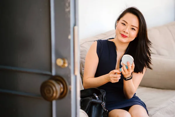 young Chinese Asian woman is applying makeup on a couch in the day next to a door. She's smiling and is looking cheerful, sunny and is professionally dressed in a dark, navy blue dress with bag