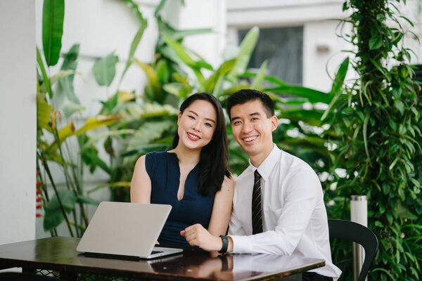 Portrait of two smiling business people (lawyers, consultants, etc) or couple smiling mid discussion over a laptop in an office with greenery in the background. They are focused on the laptop.