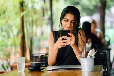 Bir kafede otururken veya smartphone ile yer coworking bir çekici, genç Hintli kadın samimi portresi. güneşli bir günde. Arka planda gür, yeşil bitkiler vardır.