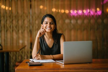 Çekici Hint Asya öğrenci profesyonel bir kafede bir masada bir kitapta yazılı. Bir dizüstü bilgisayar ve diğer sabit masasında. Kahverengi ışıkları ile arka planda.