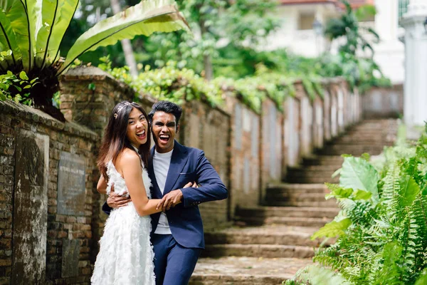 interracial couple (Indian man, Chinese woman) pose for wedding photographs and portraits in a park in the day. They are on old stone steps and and smiling and laughing as they take photos.