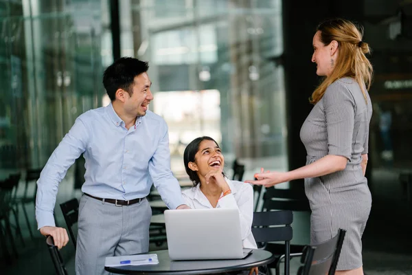 3 people from a diverse team are having a discussion around a laptop computer in the day in an open area. They are energetic and cheerful as they smile and talk.