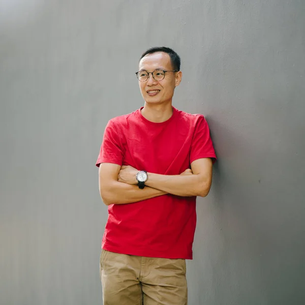 Portrait of a young Asian man (Chinese, Japanese, Korean) leaning against a grey wall and smiling. He looks reliable, dependable and relaxed.
