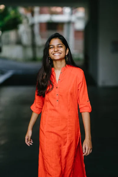 魅力的で若いインドの女性は都市の背景に笑みを浮かべてします 民族のオレンジのコスチューム ドレスを着ています — ストック写真