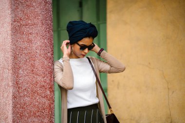 Pastel renkli duvara yaslanmış gün içinde bir güzel, genç Müslüman Arap kadın (Asya) portresi. O uzun boylu, ince ve zarif şekilde giyinmiş bir sarık, bir şık kıyafet ve güneş gözlüğü.