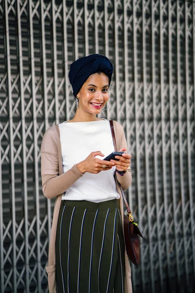 Portrait of a smartly dressed Muslim (Asian, Malay, Arab) woman wearing a turban (hijab, headscarf) talking on her smartphone. She is smartly dressed in a camisole and cardigan against a metal door.