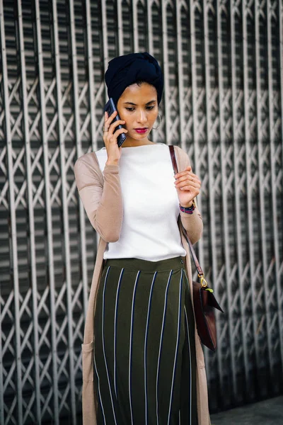 Portrait of a smartly dressed Muslim (Asian, Malay, Arab) woman wearing a turban (hijab, headscarf) talking on her smartphone. She is smartly dressed in a camisole and cardigan against a metal door.