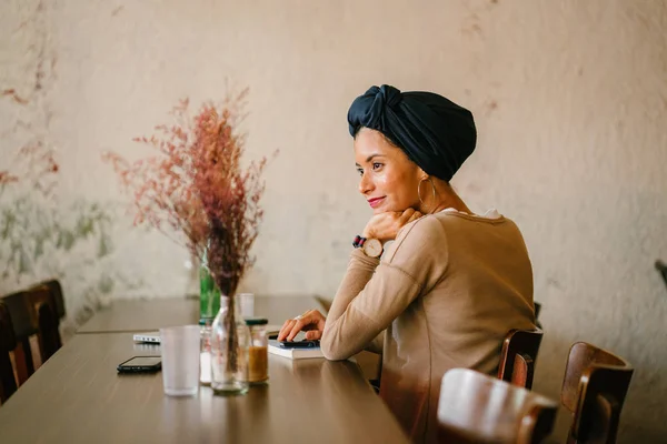 一个年轻的穆斯林妇女的肖像 她是亚洲血统 阿拉伯 马来语 和戴头巾 头围巾 并微笑着她坐在一个木桌上 她的设备花 — 图库照片