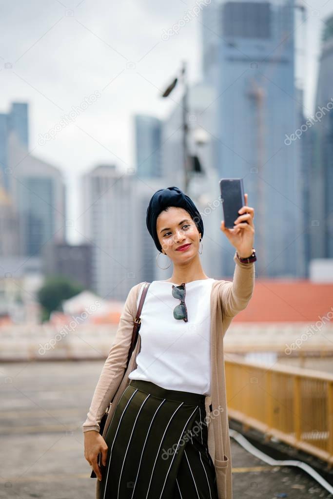 Portrait of young Muslim woman (Arab, Malay, Asian) wearing a turban (hijab, head scarf) with smartphone against a city skyline in the background (Singapore).