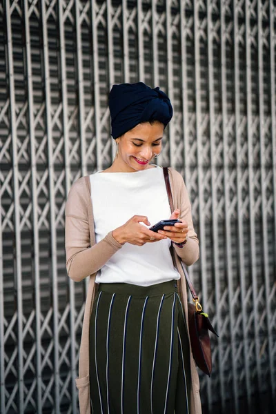 Portrait of a smartly dressed Muslim (Asian, Malay, Arab) woman wearing a turban (hijab, headscarf) with smartphone. She is smartly dressed in a camisole and cardigan against a metal door.