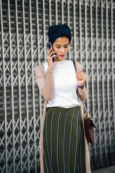 Portrait of a smartly dressed Muslim (Asian, Malay, Arab) woman wearing a turban (hijab, headscarf) with smartphone. She is smartly dressed in a camisole and cardigan against a metal door.