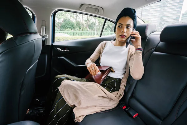Portrait of a young, attractive Muslim woman commuting in a private limousine. She is wearing a turban and smartly but modestly dressed in the backseat and talking on her smartphone.