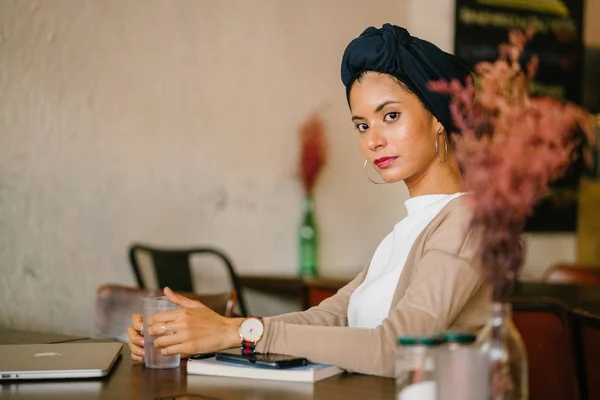 Portrait of a young Muslim woman. She is of Asian descent (Arab, Malay, Asian) and is wearing a turban (hijab, head scarf) and  she sits at a wooden table