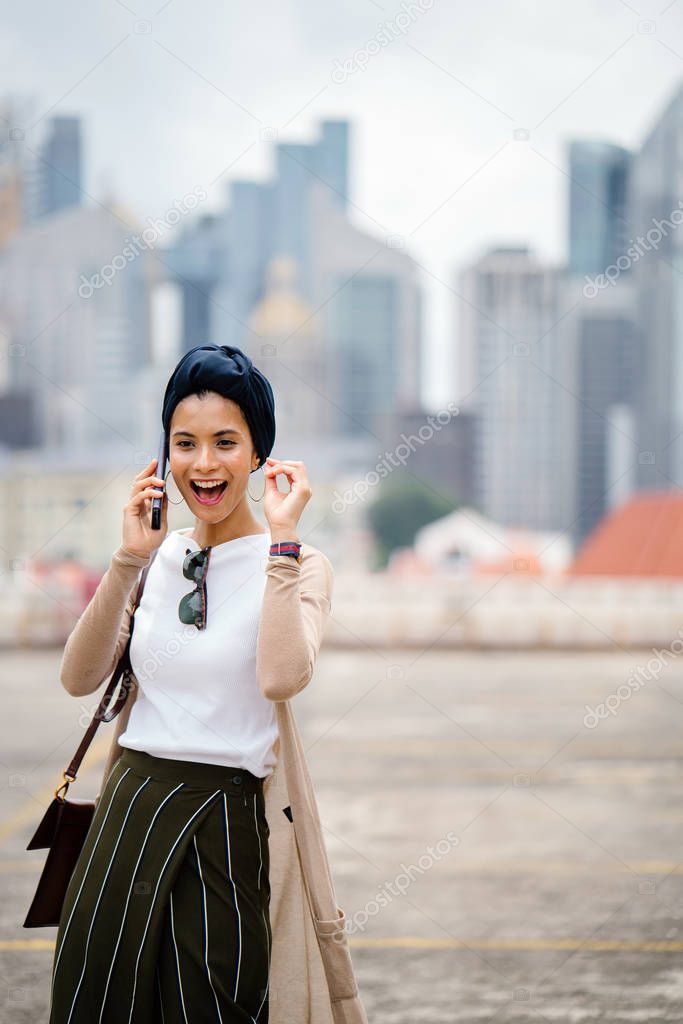 Portrait of young Muslim woman (Arab, Malay, Asian) wearing a turban (hijab, head scarf) with  smartphone against a city skyline in the background (Singapore).