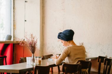 genç, çekici Müslüman kadınla (Arap, Malay, Asya) gün kafede telefon. O bir Türban (başörtüsü, hijabi) giyiyor ve dünyevi tonlarda zarif giyinmiş.