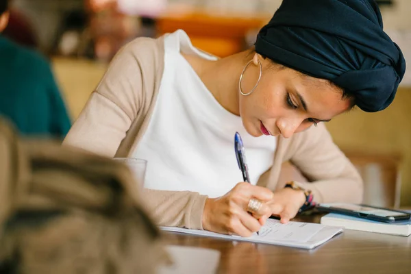 young, attractive Muslim student (Arab, Malay, Asian) writing in notepad in cafe in the day. She is wearing a turban (headscarf, hijabi) and is elegantly dressed in earthy tones.