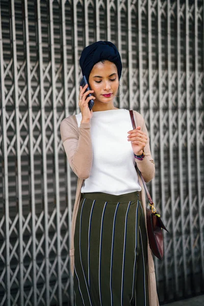 Portrait of a smartly dressed Muslim (Asian, Malay, Arab) woman wearing a turban (hijab, headscarf) with smartphone. She is smartly dressed in a camisole and cardigan against a metal door.