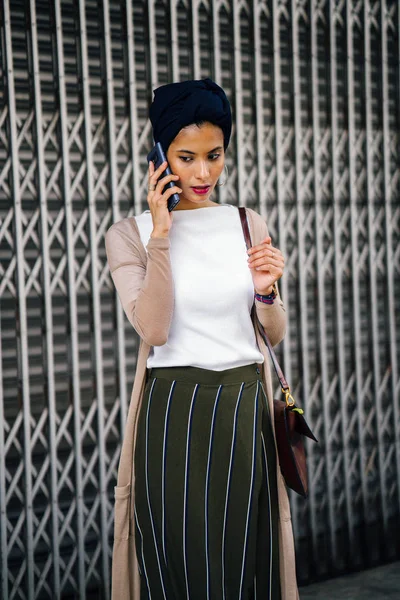 Portrait of a smartly dressed Muslim (Asian, Malay, Arab) woman wearing a turban (hijab, headscarf) with smartphone. She is smartly dressed in a camisole and cardigan against a metal door.