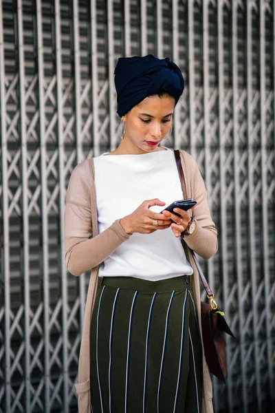 身着智能手机的穆斯林 阿拉伯 妇女戴头巾 的肖像 她穿着一件背心的毛衣 穿上一扇金属门 — 图库照片