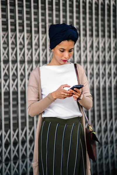 Portrait of a smartly dressed Muslim (Asian, Malay, Arab) woman wearing a turban (hijab, headscarf) with smartphone. She is smartly dressed in a camisole and cardigan against a metal door.