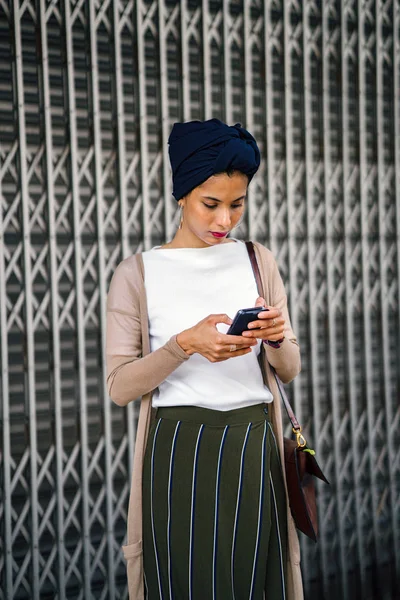 Portrait of a smartly dressed Muslim (Asian, Malay, Arab) woman wearing a turban (hijab, headscarf) with smartphone. She is smartly dressed in a camisole and cardigan against a metal door.