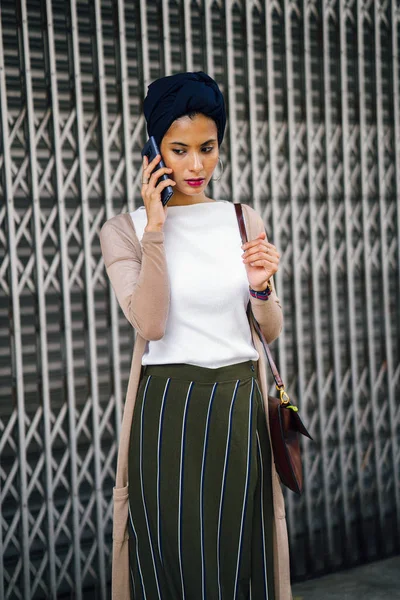 Portrait of a smartly dressed Muslim (Asian, Malay, Arab) woman wearing a turban (hijab, headscarf) with smartphone. She is smartly dressed in a camisole and cardigan against a metal door.